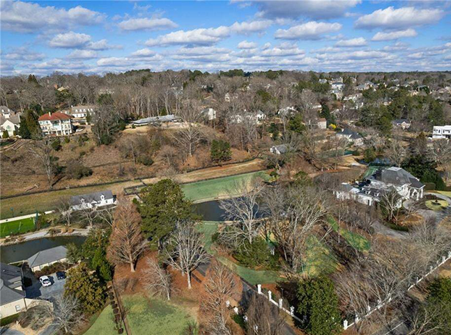 Aerial view of a suburban neighborhood with scattered houses, leafless trees, winding roads, and a small pond or creek running through the landscape under a partly cloudy sky—some homes show signs of recent ceiling repair.