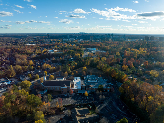 Aerial view of a suburban area with colorful autumn trees, houses, and buildings. Drywall Experts work in the neighborhood as the city skyline is visible in the distance under a blue sky with scattered clouds.