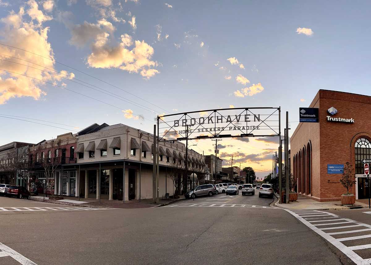 A street view of downtown Brookhaven at sunset, featuring a sign that reads “Brookhaven, Home Seekers Paradise.” Cars are parked along the street, with buildings on both sides under a partly cloudy sky—perfect for strolling after a day of popcorn removal.
