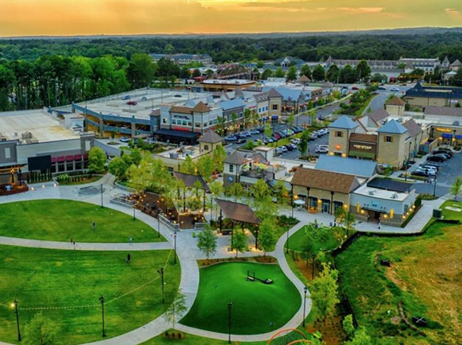 Aerial view of a modern outdoor shopping and recreational area with green lawns, walking paths, small buildings featuring ceiling renovation, trees, and parked cars, set against a sunset and wooded background.
