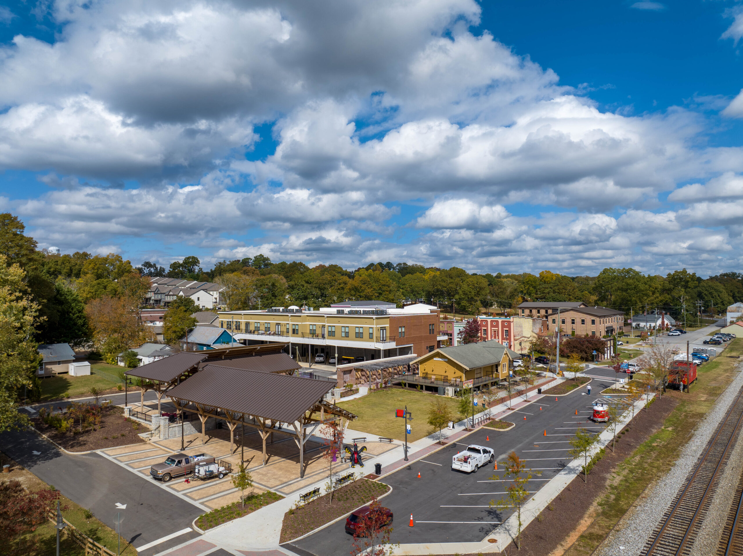 Aerial view of a small town train station featuring a covered pavilion with recent ceiling modernization, nearby buildings, parked cars, and train tracks running parallel to the road under a partly cloudy blue sky.