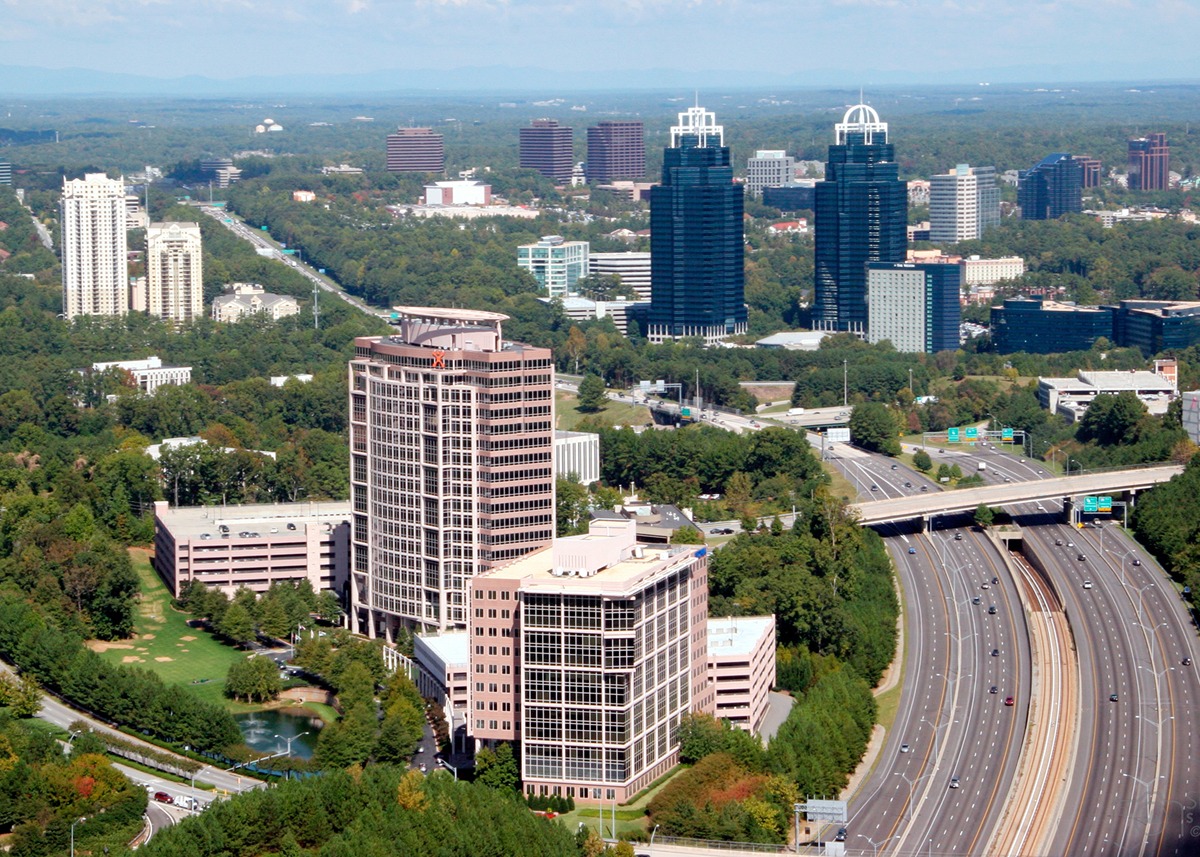Aerial view of a cityscape with tall office buildings surrounded by greenery. Popcorn removal services are available nearby as a multi-lane highway with moving cars runs alongside the buildings, and more skyscrapers and trees appear in the distance.