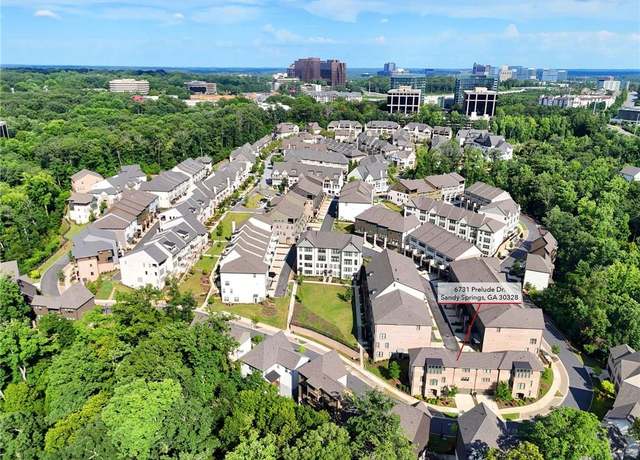 Aerial view of a residential neighborhood surrounded by trees, featuring clusters of townhouses and apartment buildings—some recently updated with popcorn removal. A city skyline is visible in the background under a clear blue sky.