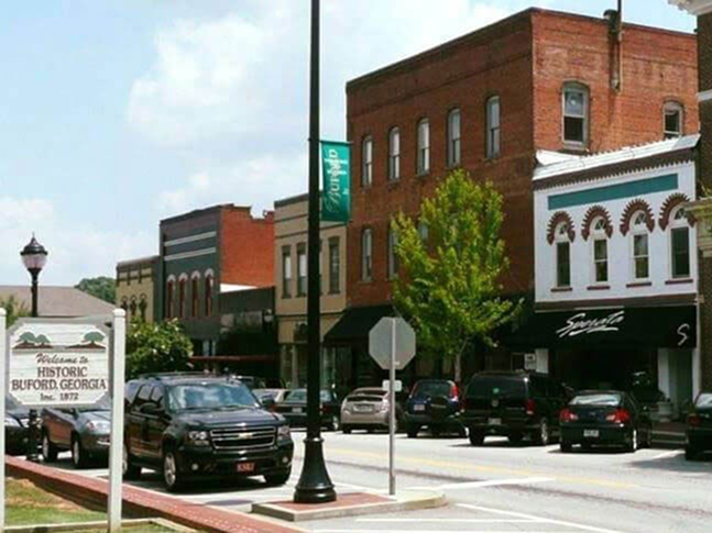 A downtown street in Buford, Georgia, with brick buildings, storefronts, parked cars, a green banner, trees, and a sign welcoming visitors to historic Buford—home to trusted local services like drywall repair. The sky is partly cloudy.