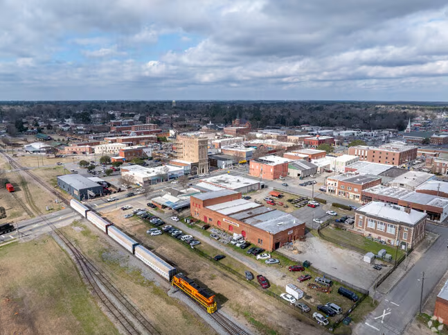 Aerial view of a small town with brick buildings, parking lots, Drywall Experts signage, and a yellow and orange train traveling on railroad tracks under a partly cloudy sky.