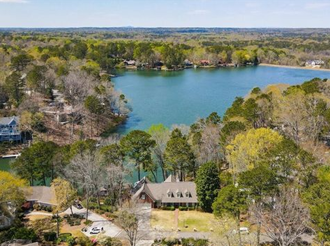 Aerial view of a lake surrounded by trees and houses, with one large house—perfect for drywall experts—prominently visible in the foreground and more homes scattered along the shoreline in the background.