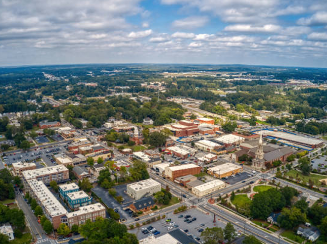 Aerial view of a small town with a mix of historic and modern buildings, roads, trees, and a church with a tall steeple under a partly cloudy sky. Local businesses include home services such as expert drywall repair amid the green, sprawling landscape.