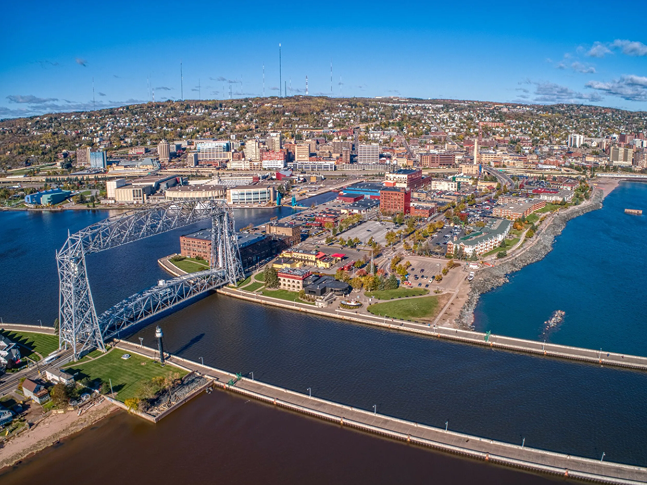 Aerial view of a city by a large lake, featuring a distinctive vertical-lift bridge, waterfront parks, and a downtown area with various buildings—some showcasing recent drywall repair—under a blue sky.