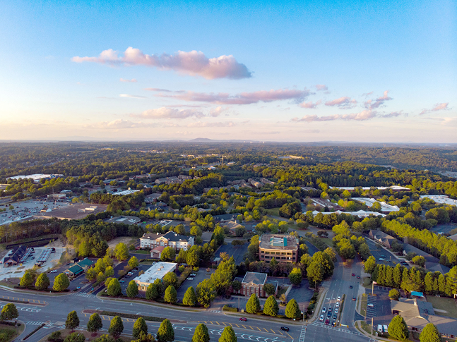 Aerial view of a suburban area with tree-lined streets, commercial buildings, parking lots, and dense green forests under a blue sky. Roads and intersections are visible, including businesses specializing in ceiling renovation.