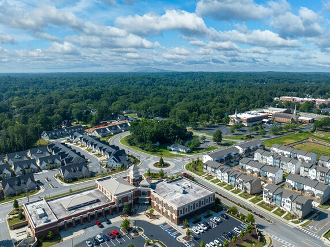 Aerial view of a suburban neighborhood with rows of houses and wide streets, featuring a large building—possibly undergoing ceiling renovation—in the foreground, all surrounded by dense green trees under a partly cloudy sky.