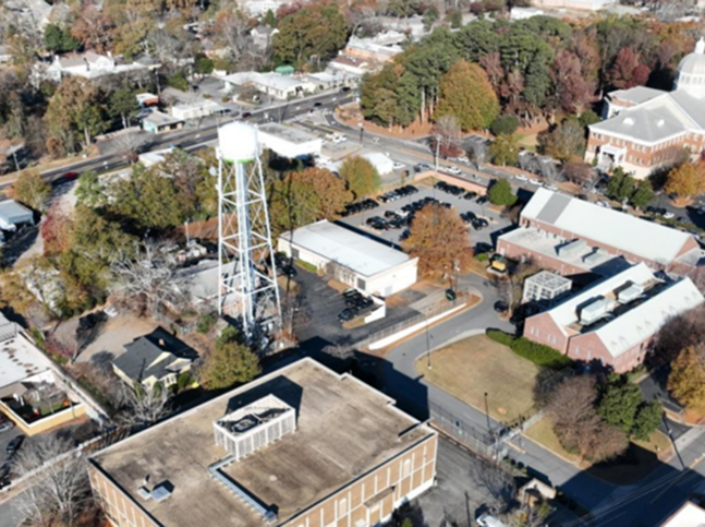 Aerial view of a small town area with a white water tower, several brick and light-colored buildings, parking lots, roads, trees with autumn foliage, and signs of recent ceiling repair on some rooftops.