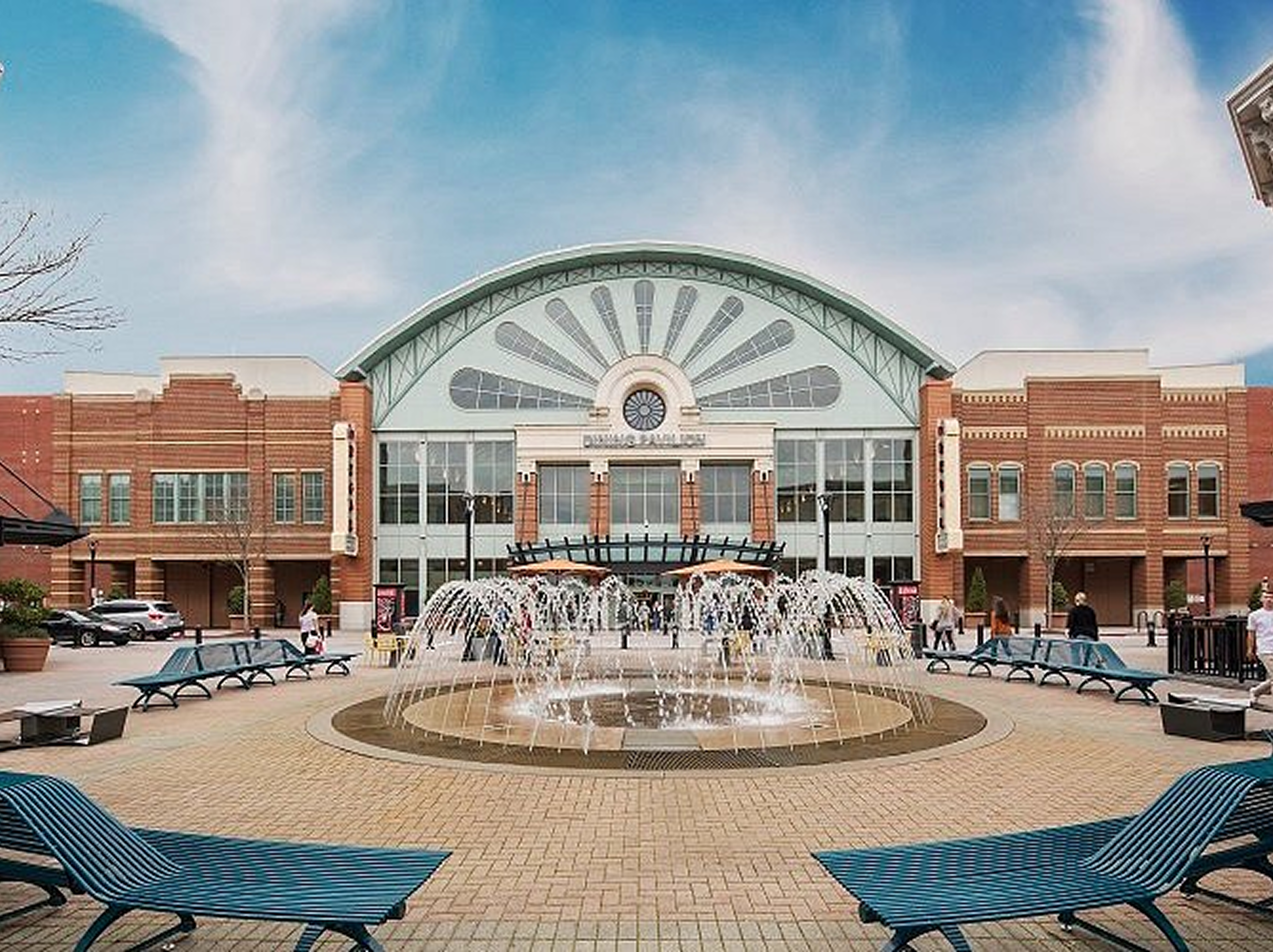 A large shopping mall entrance with an arched glass facade showcases recent ceiling modernization. A prominent clock, surrounded by brick buildings, overlooks a circular fountain and blue benches in the foreground, as people walk nearby under a blue sky.